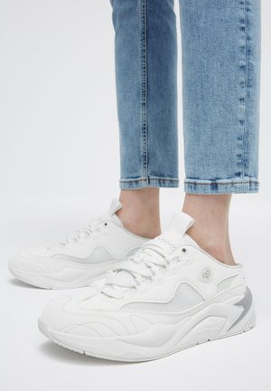 Feet wearing white slip-on sneakers with laces and light blue jeans cropped above the ankle on a white background.