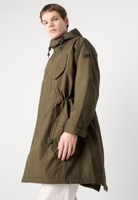 Parka impermeável verde-oliva com cintura ajustável por cordão, grandes bolsos frontais, capuz ajustável e botões de pressão para fecho.