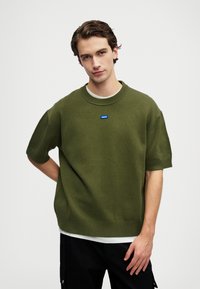 Πράσινο πλεκτό πουλόβερ με κοντά μανίκια και στρογγυλή λαιμόκοψη. Διαθέτει μπλε λογότυπο στο στήθος και ριγωτές μανσέτες και στρίφωμα. Φορεμένο πάνω από μια λευκή μπλούζα.