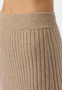Gros plan sur la taille portant un pantalon beige en maille côtelée avec une large ceinture élastique, sur un fond neutre.