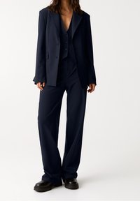 Costume trois pièces bleu marine à fines rayures, comprenant un long blazer, un gilet et un pantalon à jambes larges avec une coupe décontractée et des détails à boutons.