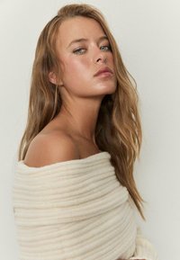 Mehak, kremno obarvan pulover z rebrasto teksturo, ovit čez ramena, razkriva golo kožo na enem rami in dolge, valovite lase.