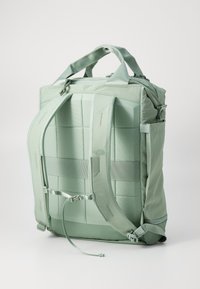 Mochila de tela verde con respaldo acolchado y correas ajustables, que presenta un diseño minimalista, clips laterales y un patrón de costura en cuadrícula.