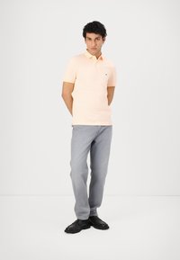 Lyse ferskenfarvet polo T-shirt med krave og lille logo, parret med lysegrå jeans og sorte sko. Simpelt design og glat tekstur.