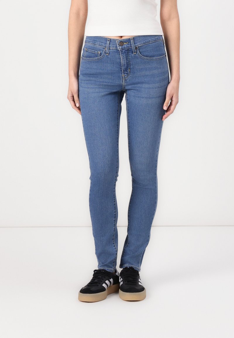 Jean bleu skinny avec taille mi-haute, cinq poches et un détail cousu. Associé à des baskets noires avec des rayures grises.