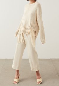 Beige neulepaita, jossa on fringe-yksityiskohtia helmassa, yhdistettynä löysiin kermaisiin housuihin ja vaaleankeltaisiin avokärkisiin kenkiin.
