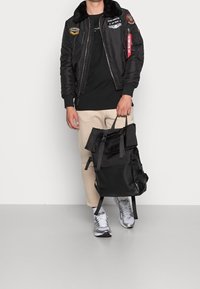 Černá bomber bunda s nášivkami, béžové kalhoty a černý látkový batoh. Stříbrné tenisky se šedými akcenty. Jednoduchý, neformální design.