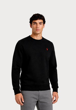 Man wearing a black crewneck sweater with a small red embroidered logo on the chest and gray pants standing against a plain background.