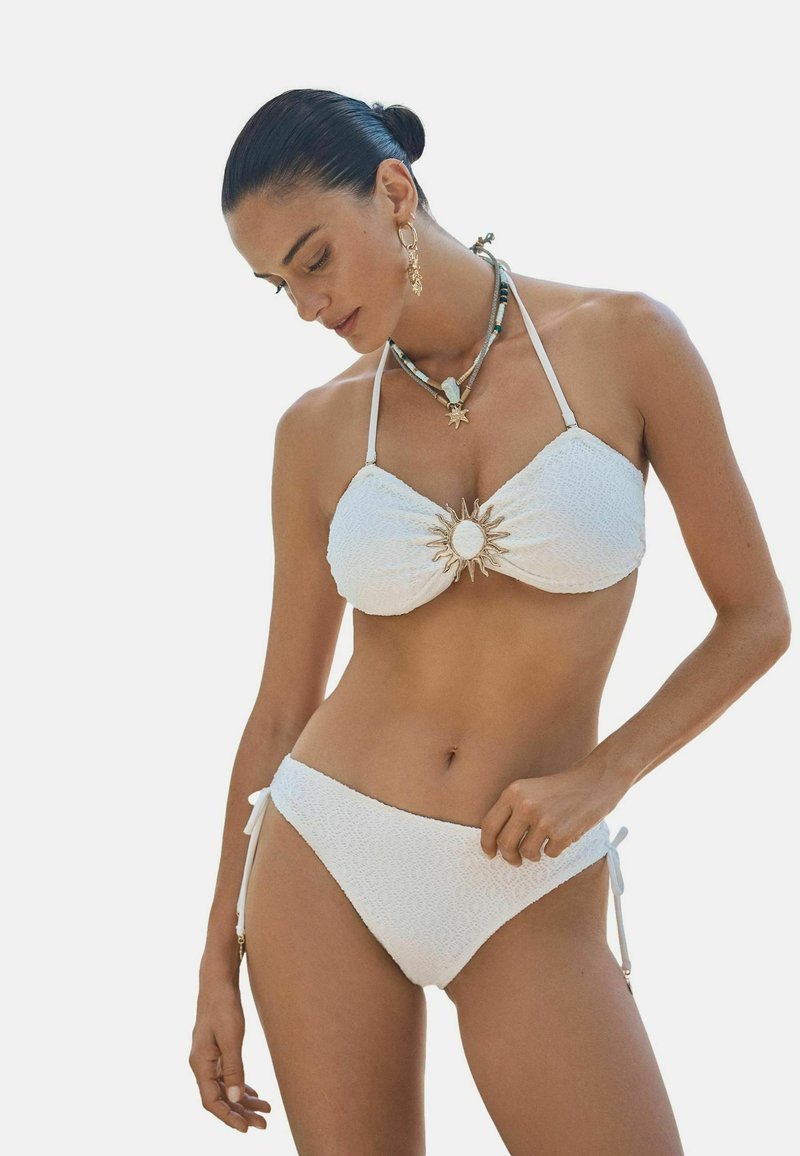 Tumejuustega naine valges tekstuurse bikiini ülaosaga, millel päikesekujuline kaunistus, kannab kihtides kaelakeesid ja kuldseid kõrvarõngaid, vaadates alla.