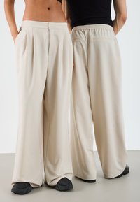 Pantalons larges beiges avec des plis à l'avant, des poches latérales et une taille élastique à l'arrière. Le tissu semble lisse avec un léger éclat.