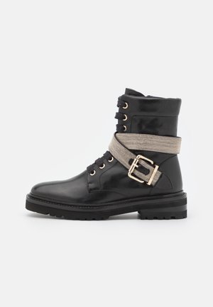 Czarny skórzany botek za kostkę z grubą podeszwą, sznurowany z przodu, z szerokim metalicznym paskiem z złotą klamrą wokół kostki.