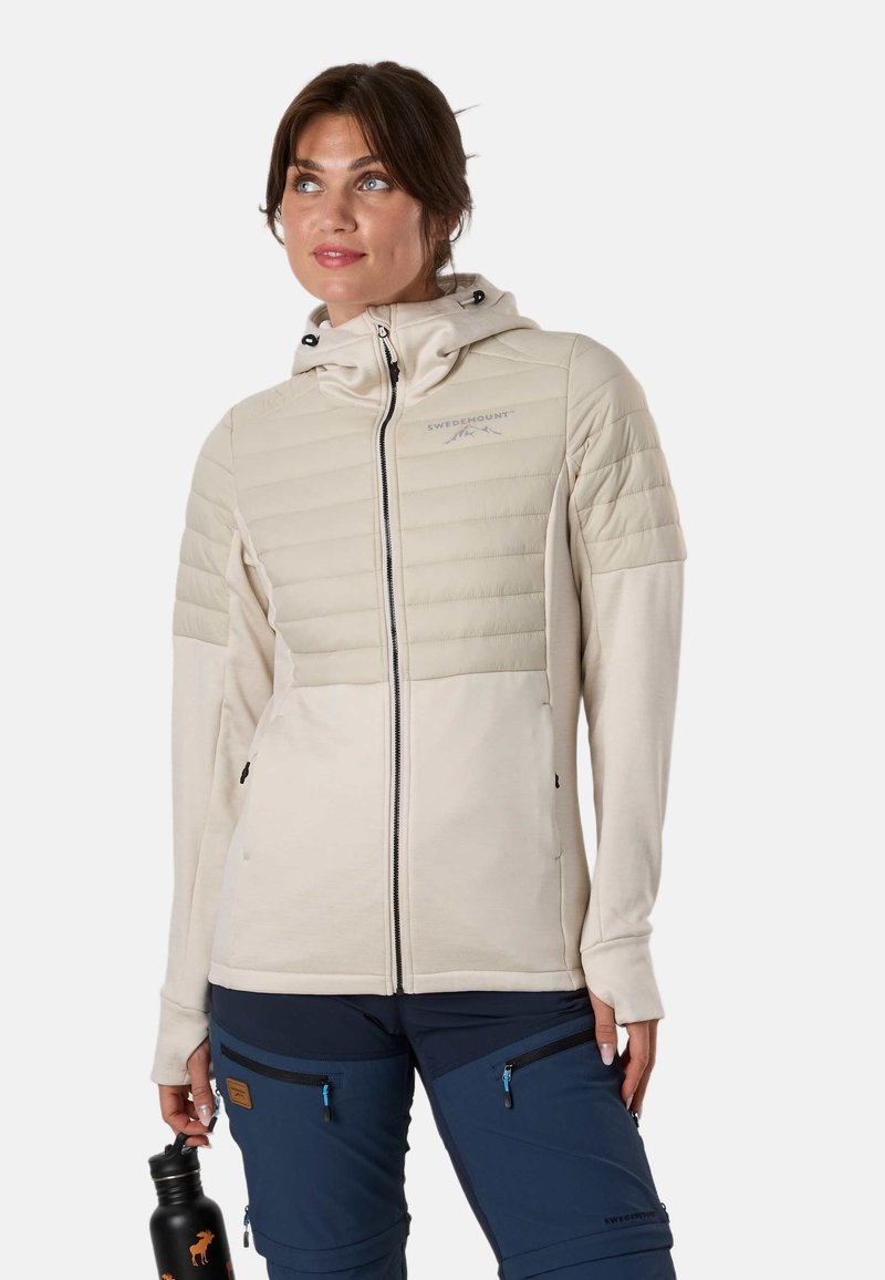 Femme portant une veste matelassée beige et un pantalon de plein air bleu marine, tenant une bouteille d'eau noire avec des logos d'élans.