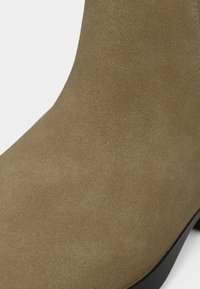 Hnědá semišová kotníková botka, texturovaný povrch, zaoblený tvar, černá gumová podrážka, minimální kovové prvky, elegantní design, bezproblémové švy.