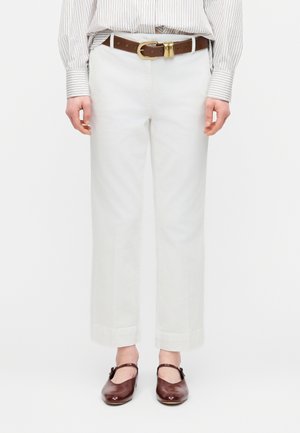 Pantaloni bianchi a gamba larga tagliati sopra la caviglia con orli risvoltati, abbinati a una cintura marrone e scarpe Mary Jane marrone scuro su una persona.