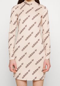Lichtroze lange mouwen jurk met een col, met herhalende bruine Adidas-logo's en trefoil-patronen op een glanzende, gestructureerde stof.