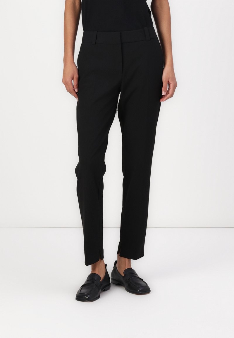 Personne portant un pantalon noir droit avec de petites fentes à l'avant au niveau de l'ourlet et des mocassins en cuir noir, debout sur un fond blanc.