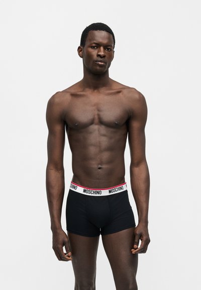 Hombre atlético de piel oscura de pie, con bóxer Moschino negros con cintura blanca y ribete rojo, frente a un fondo blanco liso.