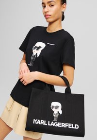 Black tote bag with cartoon character design, featuring text "KARL LAGERFELD" in bold white. Accompanied by a black t-shirt with similar design.