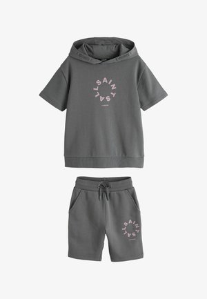 Fiú áll részben zsebre tett kézzel, szürke kapucnis pulóverben és rövidnadrág szettben, rajta rózsaszín kör alakú szöveg, fehér zokniban és fehér tornacipőben.