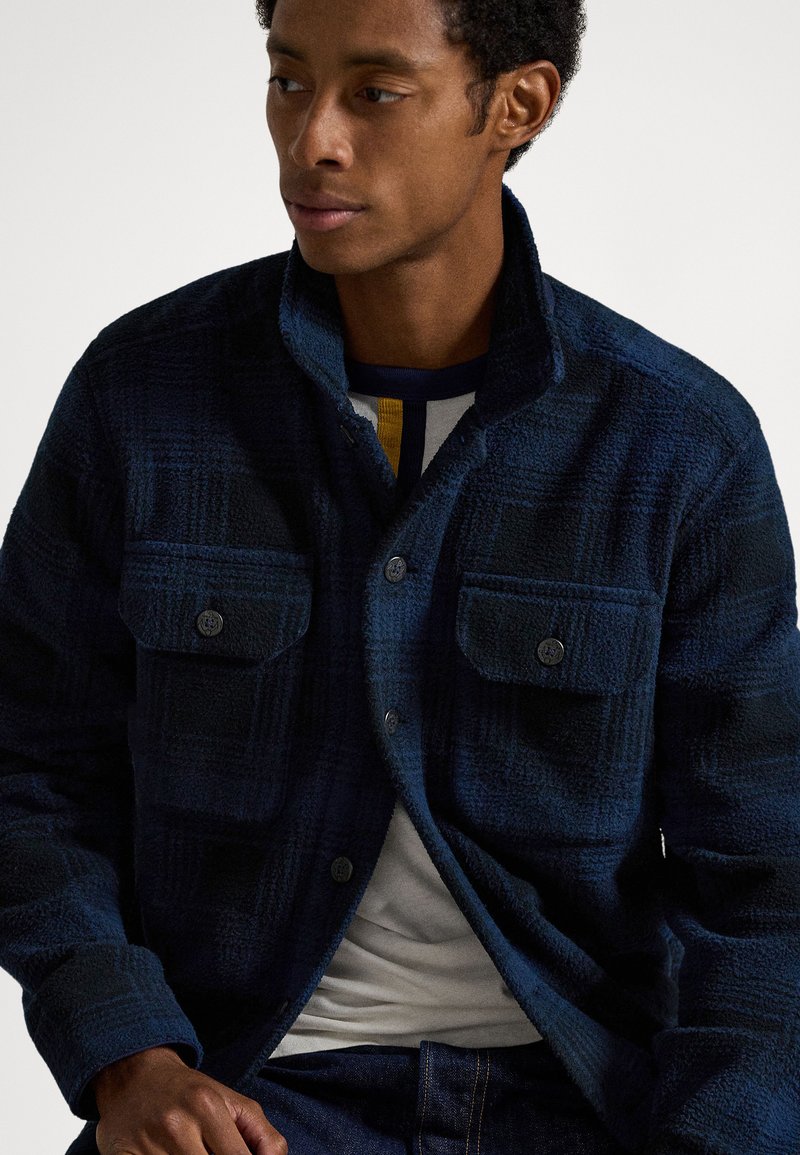 Mann trägt eine marineblaue karierte Fleecejacke mit Knöpfen über einem weißen Hemd mit gestreiftem Kragen, sitzt mit den Händen auf den Beinen.