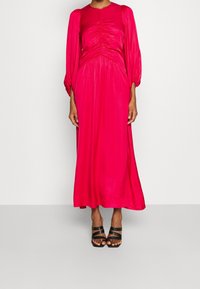 Kvinna som bär en ljusrosa långärmad midiklänning med rynkad bodice och svarta remsandaler med klack framför en neutral bakgrund.