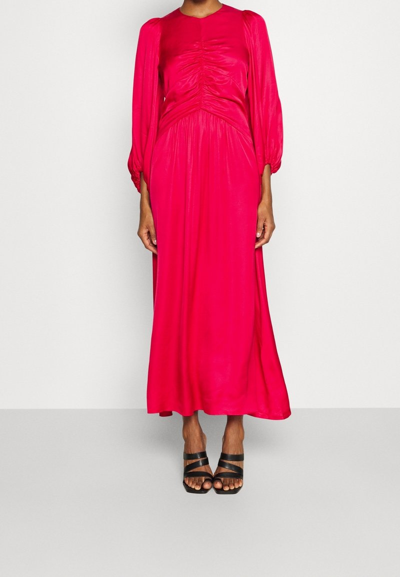 Kvinna som bär en ljusrosa långärmad midiklänning med rynkad bodice och svarta remsandaler med klack framför en neutral bakgrund.