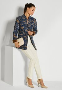 Vrouw leunt op een witte blok en draagt een blauw gemusterd jack, witte broek, beige hakken, en houdt een beige clutch vast.