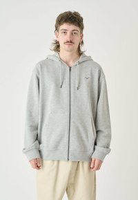 Grau zip-up Hoodie aus weichem Material, mit einer Kängurutasche, verstellbarem Kordelzug und subtilen Logo-Details auf der Brust.