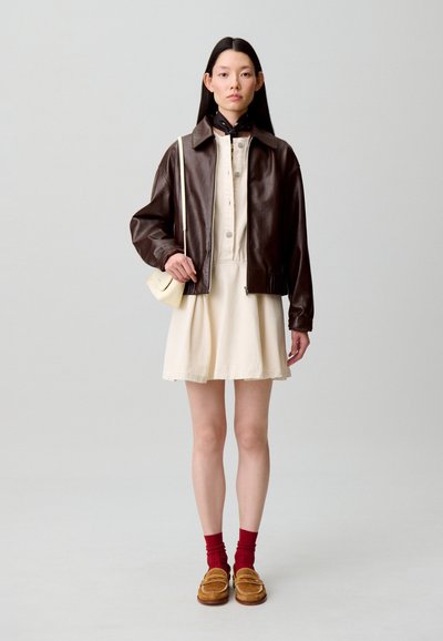Chaqueta bomber de cuero marrón sobre un vestido crema, combinada con calcetines rojos y zapatos tipo mocasín en tono beige. La modelo lleva un bolso pequeño en color blanco roto.