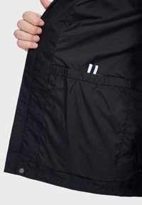 Intérieur de veste noire imperméable avec une texture lisse, mettant en avant une poche cachée avec un accent rayé bleu et blanc. Fermeture à pression simple.