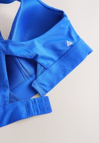 Sinine spordirinnahoidja, millel on sile kangas, võrgust detailid, reguleeritavad konksud ja väike logo paelal.