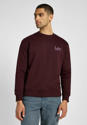 Mężczyzna zwrócony w lewo, ubrany w bordową bluzę Lee i niebieskie dżinsy, na tle gładkiej jasnoszarej ściany.