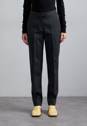 Person wearing tailored black trousers and woven beige heeled loafers, standing on a gray floor against a plain light wall.