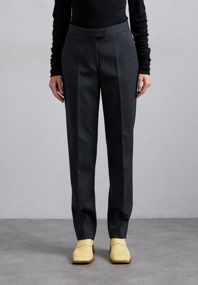 Personne portant un pantalon noir bien ajusté et des mocassins à talons tressés beige, se tenant sur un sol gris contre un mur clair et unicolore.