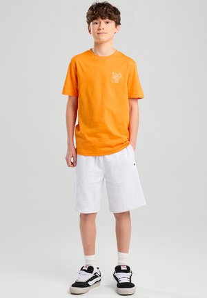 Camiseta de algodón naranja con un gráfico blanco en el pecho, combinada con pantalones cortos grises y zapatillas negras, de pie contra un fondo neutro.