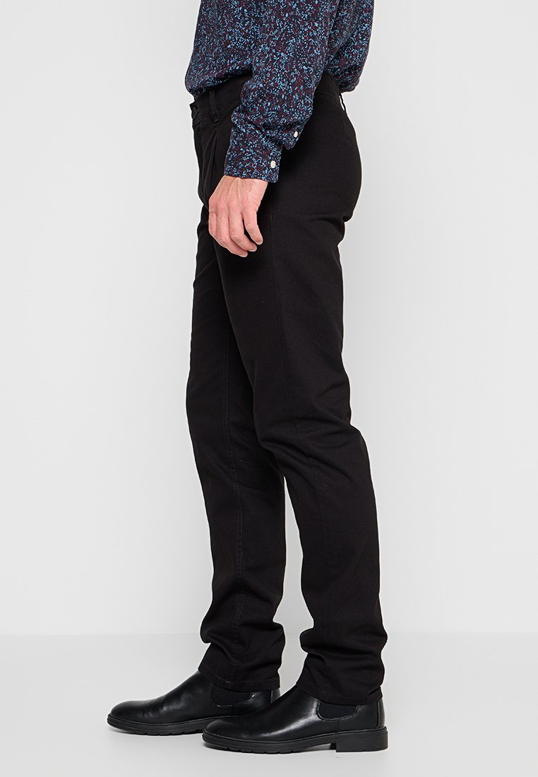 Mann in schwarzen, taillierten Hosen, schwarzen, ledernen Slippers und einem langärmeligen, gemusterten blauen Hemd, der seitlich steht.