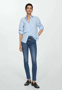 Plava i bijela prugasta košulja s dugim rukavima i podvrnutim rukavima, u kombinaciji s tamnoplavim uskim trapericama i crnim cipelama na vezivanje.