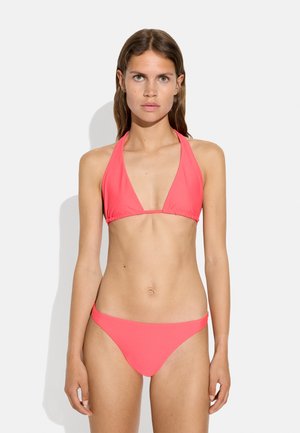 Kobieta o długich brązowych włosach, ubrana w różowy bikini halter, stojąca na gładkim białym tle, zwrócona twarzą do przodu z neutralnym wyrazem.