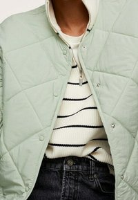 Lyst grøn, quiltet jakke med højt krave og trykknapper, båret over en hvid og marineblå stribet sweater med ribbet tekstur.