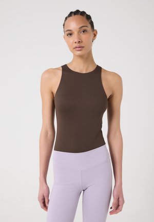 Jeune femme aux cheveux tressés portant un haut marron sans manches et des leggings taille haute violet clair, debout devant un fond uni.