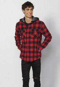 Piros és fekete kockás flannel ing kapucnival, két mellzsebbel és gombos záródással. Puha anyag, laza szabással. Fekete farmernadrág viselve alatta.