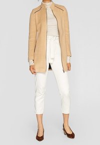Beige long coat with a high collar and horizontal zipper, paired with white cropped pants and brown leopard print heels.