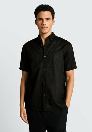 Mężczyzna z kręconymi włosami, ubrany w czarną koszulę na krótki rękaw z guzikami, stojący na jednolitym jasnoszarym tle.