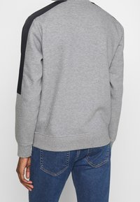 Felpa grigia con accenti neri sulle spalle, polsini e orlo ribattuti, tessuto morbido, design con girocollo, vestibilità rilassata, indossata con jeans in denim blu.