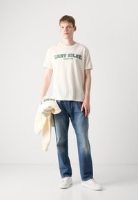 Cream-colored t-shirt with green "GANT NH. CT." text, paired with blue jeans. A matching cream sweatshirt is held in the left hand.