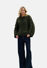 Ciemnozielony sweter z grubym splotem i oversize'owymi rękawami, zestawiony z szerokimi ciemnoniebieskimi jeansami. Sweter ma fakturowane wzory na całej powierzchni.