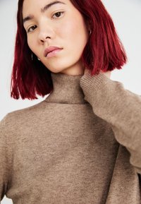 Bruin geribbelde turtleneck trui met een aansluitend ontwerp. Het model heeft kort, rood haar en raakt zachtjes haar nek aan.