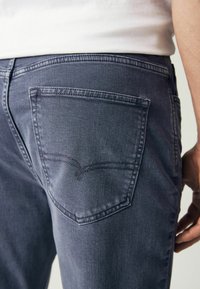 Vue arrière d'une personne portant un jean en denim bleu foncé et une chemise blanche, avec un accent sur la poche arrière droite et la zone de la hanche.