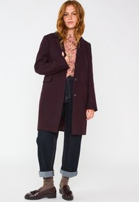 Cappotto di lana viola scuro con design sartoriale, due tasche frontali e chiusura con bottone a una fila, abbinato a jeans scuri con risvolto e scarpe.