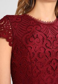 Žena oblečená v bordovém krajkovém šatu s propracovanými květinovými a spirálovými vzory, s krátkými zvlněnými rukávy a kulatým výstřihem.